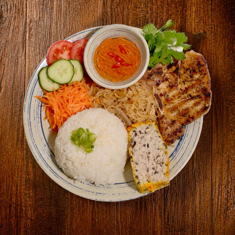 Vietnamese grilled pork plate with rice, egg loaf, pickled vegetables, cucumber, tomatoes, herbs, and dipping sauce on a wooden table.