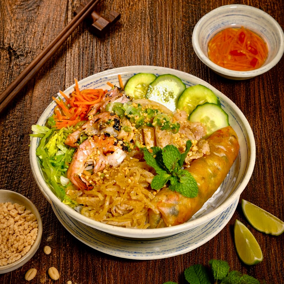 Vietnamese rice noodle bowl with shrimp, pork, spring rolls, vegetables, herbs, lime wedges, dipping sauce, and chopsticks on a wooden table.