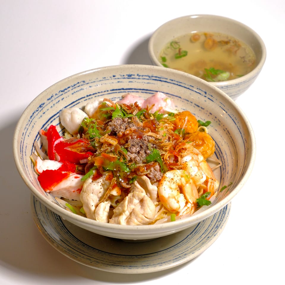 Bowl of noodles topped with shrimp, chicken, fish cakes, herbs, fried shallots, and chili, accompanied by clear soup.