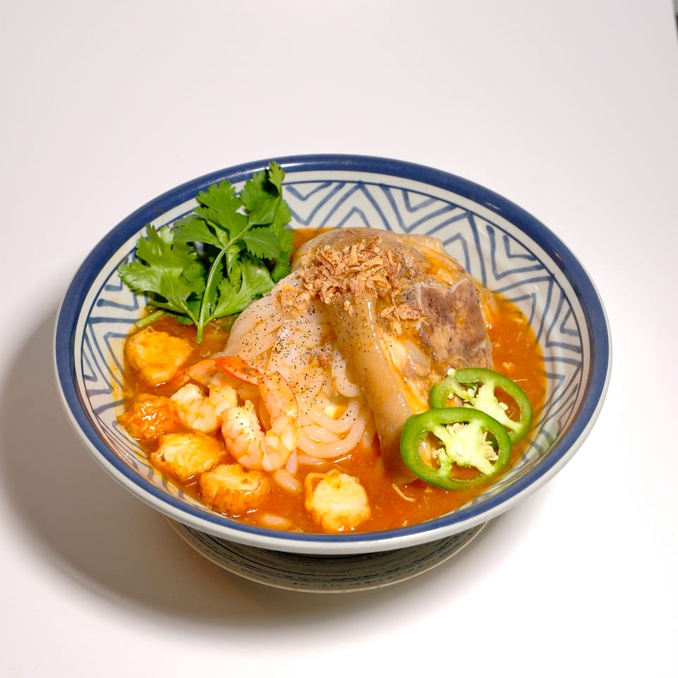 Bowl of spicy seafood soup with shrimp, fish balls, noodles, cilantro, jalapeño slices, and crispy shallots.