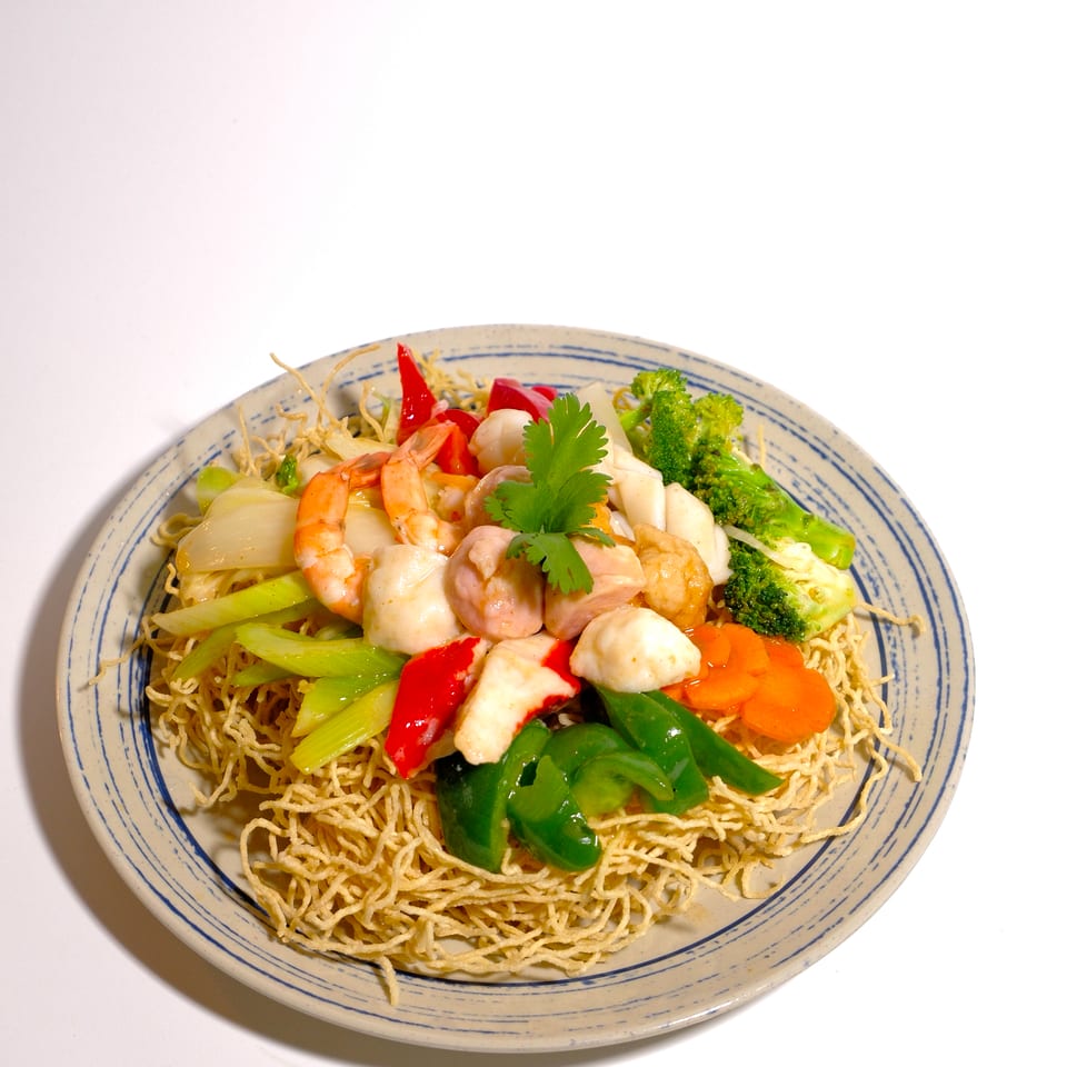 Crispy noodles topped with shrimp, chicken, seafood, broccoli, peppers, carrots, and celery on a blue-rimmed plate.