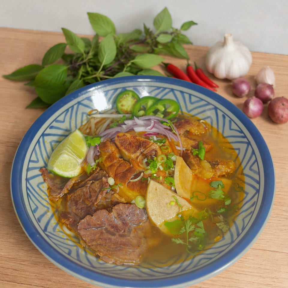 A bowl of Vietnamese-style noodle soup with beef, pork, lime, herbs, sliced chilies, and vegetables, surrounded by fresh aromatics.